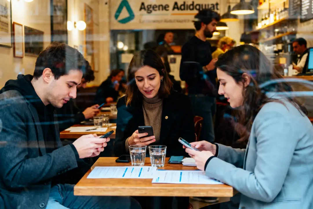 Trois personnes assises à une table, utilisant des smartphones dans un café animé.