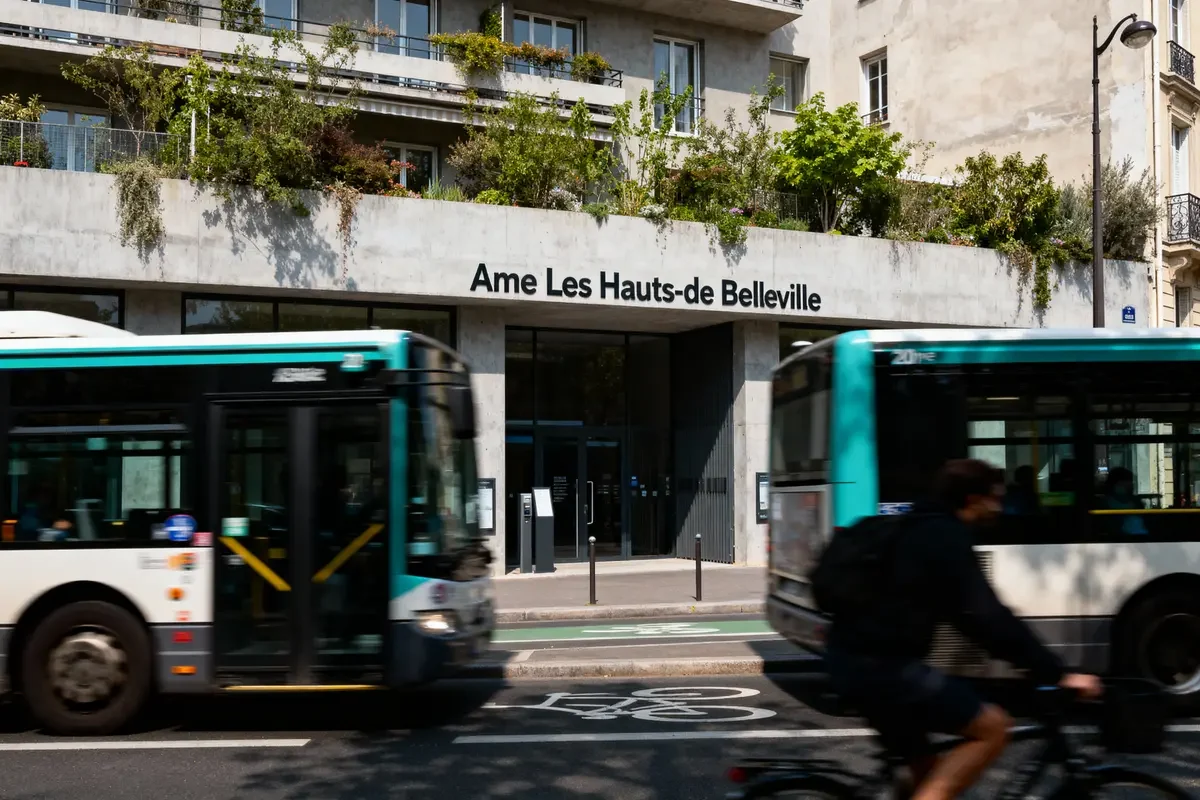 Deux bus et un cycliste passent devant l'entrée du bâtiment "Ame Les Hauts-de Belleville".
