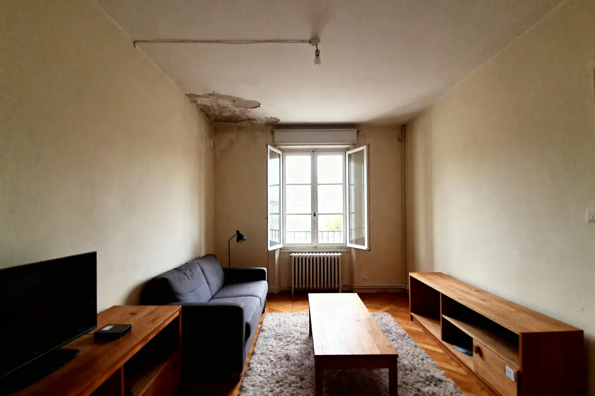 Salon simple avec canapé gris, table basse en bois, mur abîmé, fenêtre ouverte, éclairage naturel, télévision.