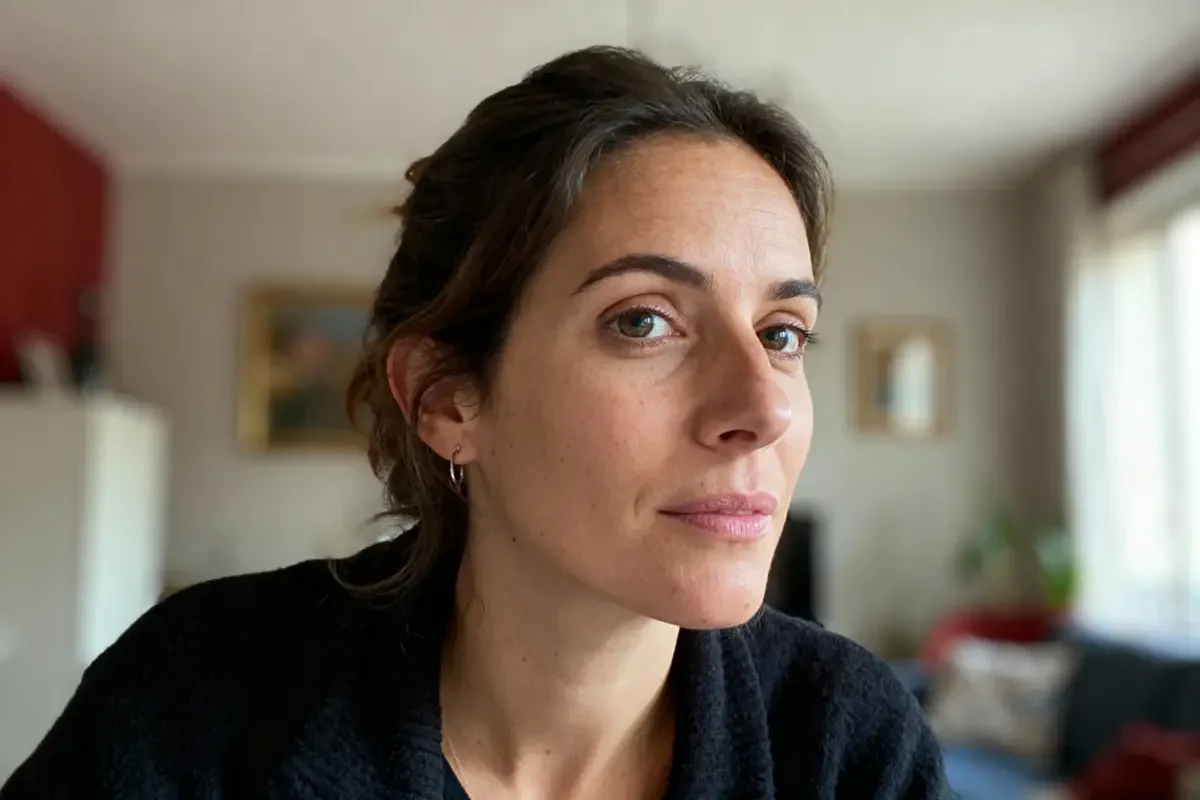 Une femme aux cheveux bruns, regarde vers le côté, fond intérieur flou, vêtements noirs, expression sereine.