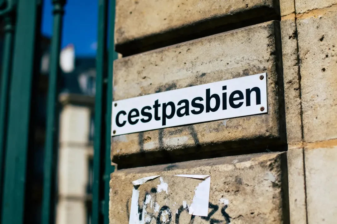 Plaque blanche avec texte "cestpasbien" fixée sur un mur en pierre. Graffiti en dessous.