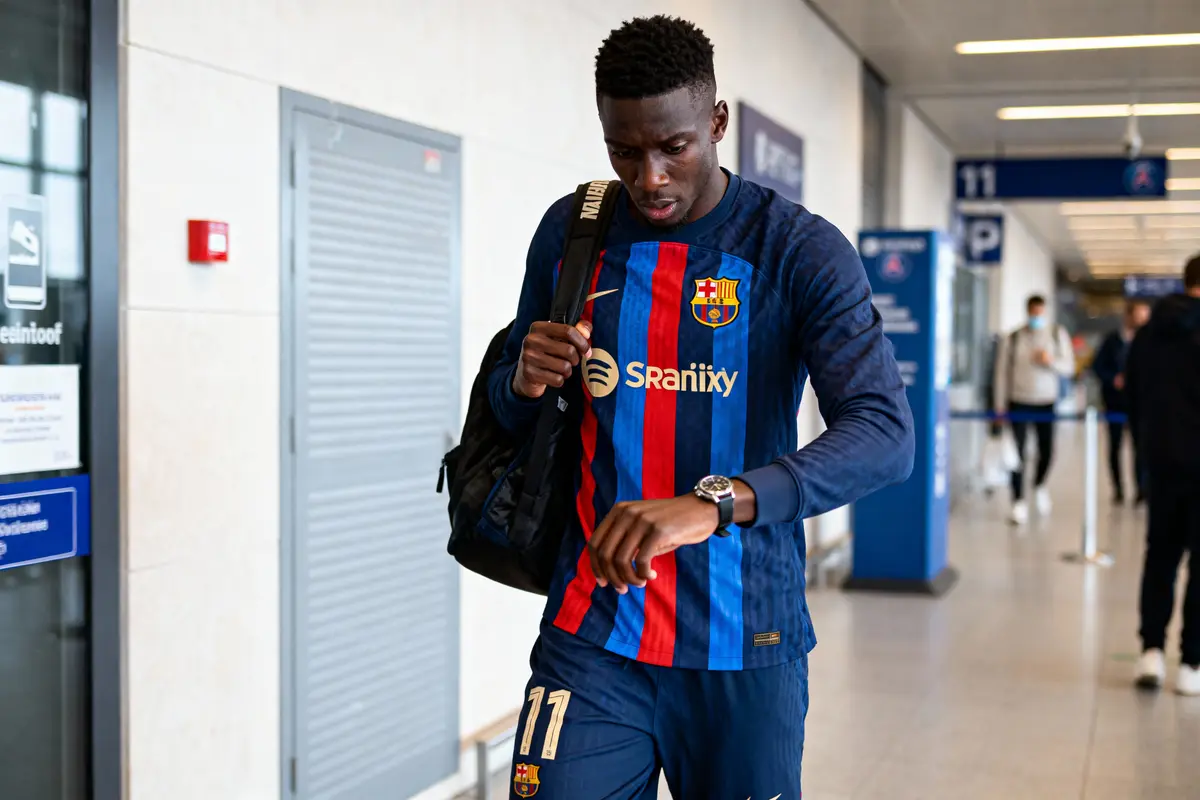 Joueur de football en maillot Barcelone vérifiant son montre dans un aéroport, tenant un sac.