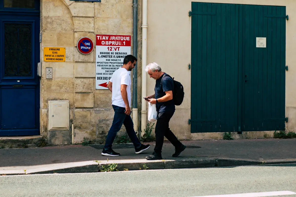 Deux personnes marchant sur un trottoir devant un mur avec panneaux, une porte bleue et volets verts.