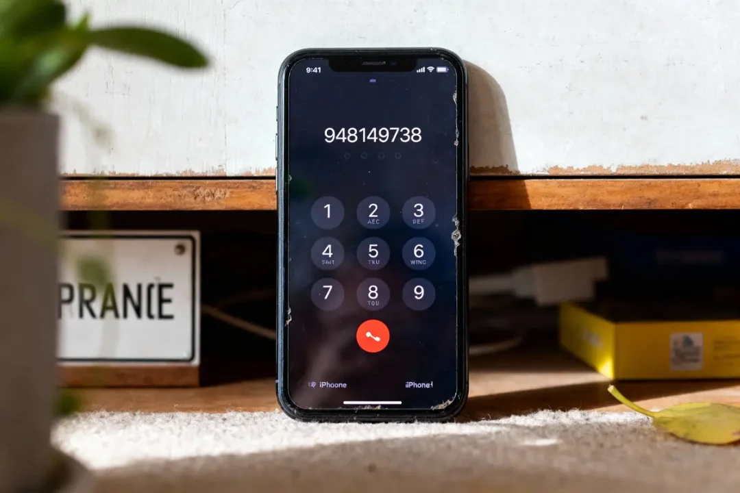 iPhone avec écran d'appel affichant un numéro, posé contre un meuble en bois, éclairé par le soleil.