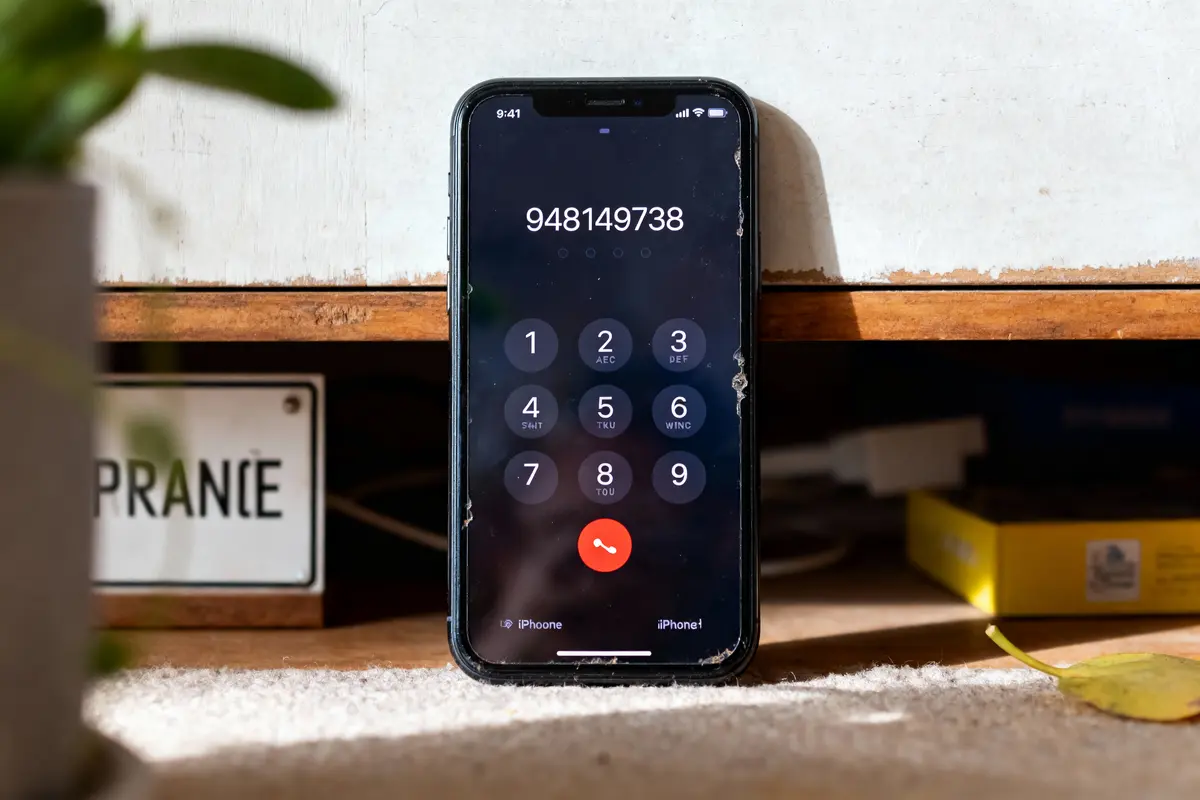 iPhone avec écran d'appel affichant un numéro, posé contre un meuble en bois, éclairé par le soleil.