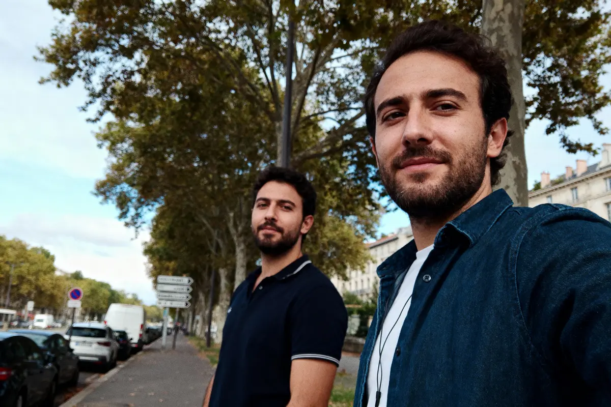 Deux hommes souriants marchant sur un trottoir bordé d'arbres en ville, ciel partiellement nuageux.