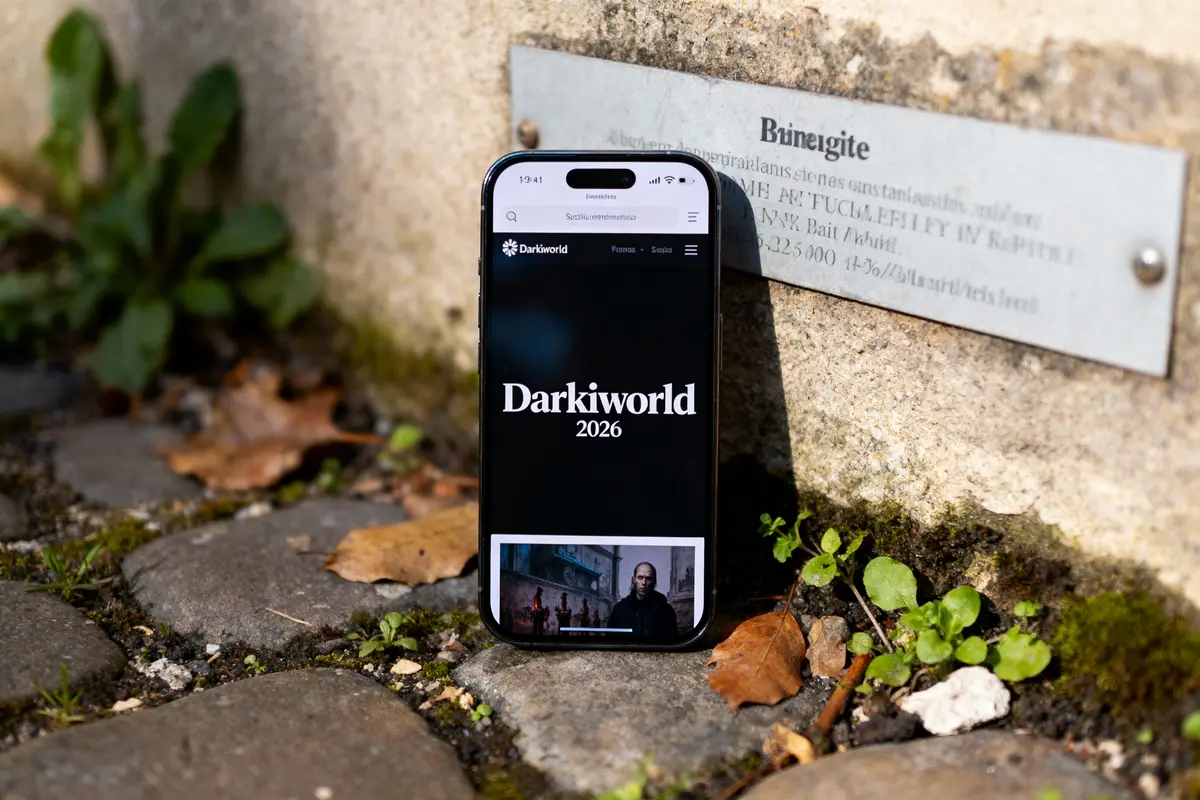 Smartphone affichant "Darkiworld 2026" sur pavés, près d'un mur en pierre avec plaque métallique et feuilles mortes.