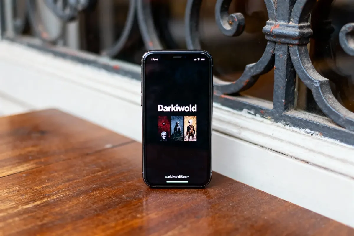 Smartphone sur table avec écran affichant "Darkiwold" et illustrations, près de garde-corps en fer forgé.