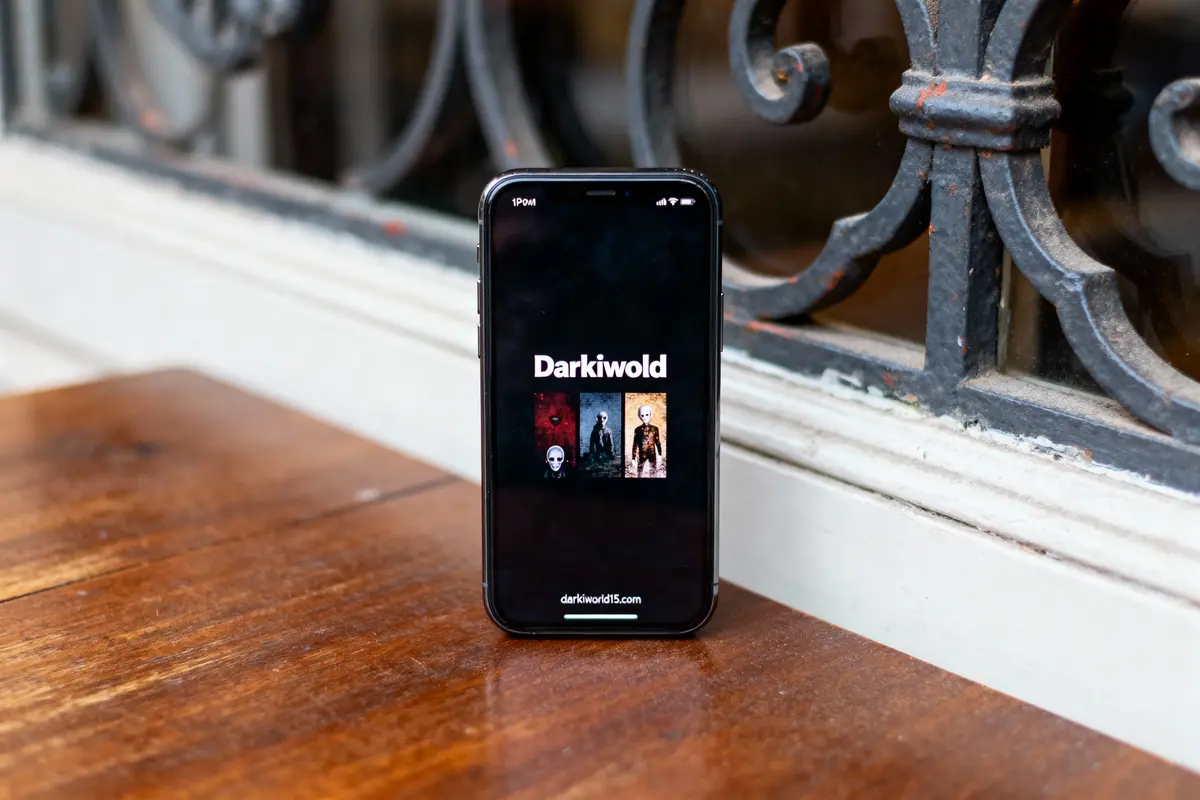 Smartphone sur table avec écran affichant "Darkiwold" et illustrations, près de garde-corps en fer forgé.