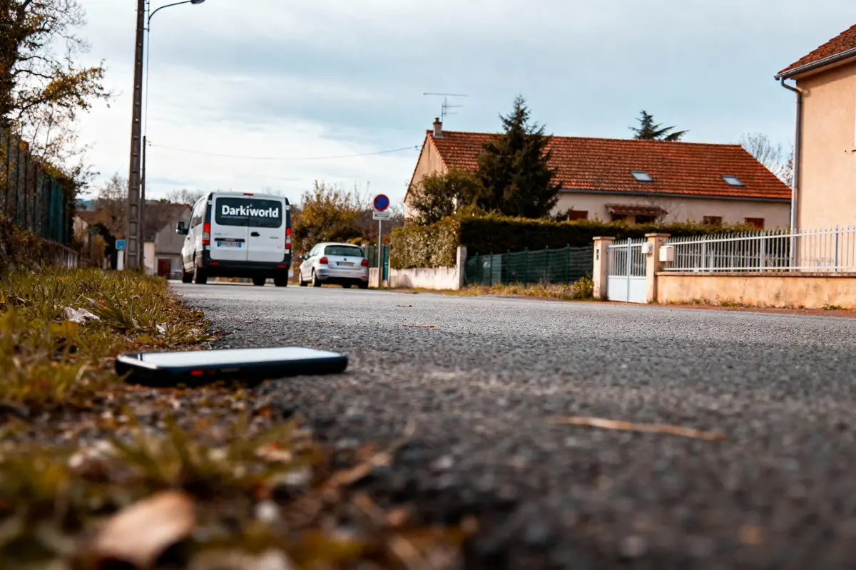 Smartphone abandonné sur route, voiture blanche garée devant maison rouge, fourgonnette "Darkiworld" en arrière-plan.