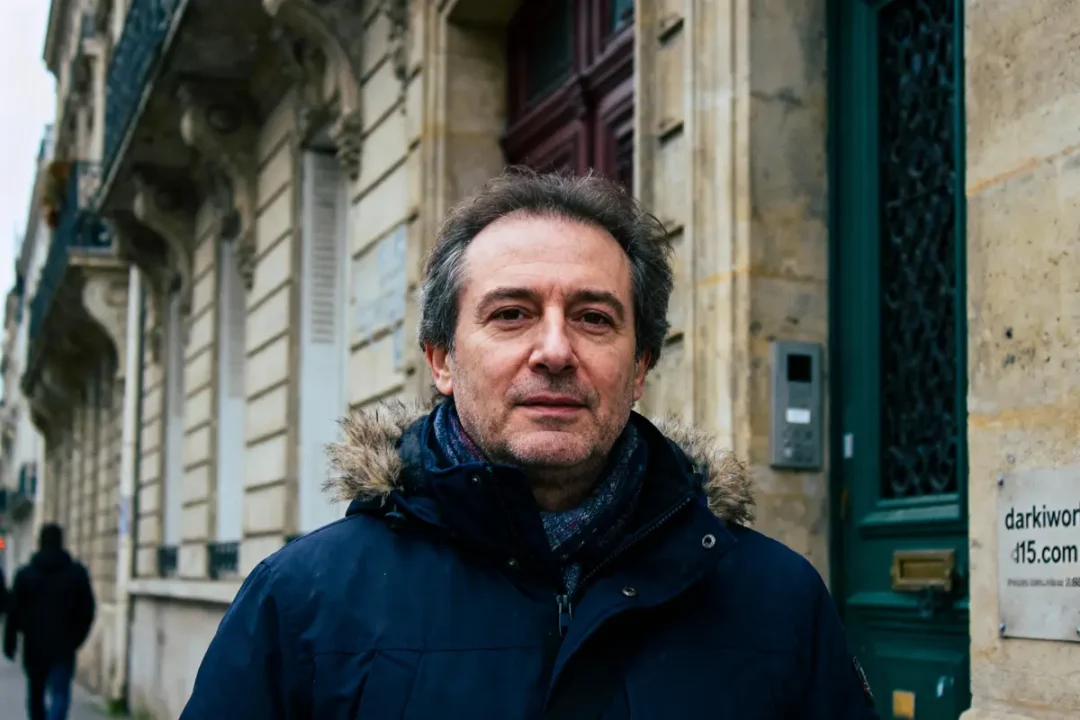 Un homme en manteau sombre se tient devant un bâtiment ancien, rue parisienne en arrière-plan.