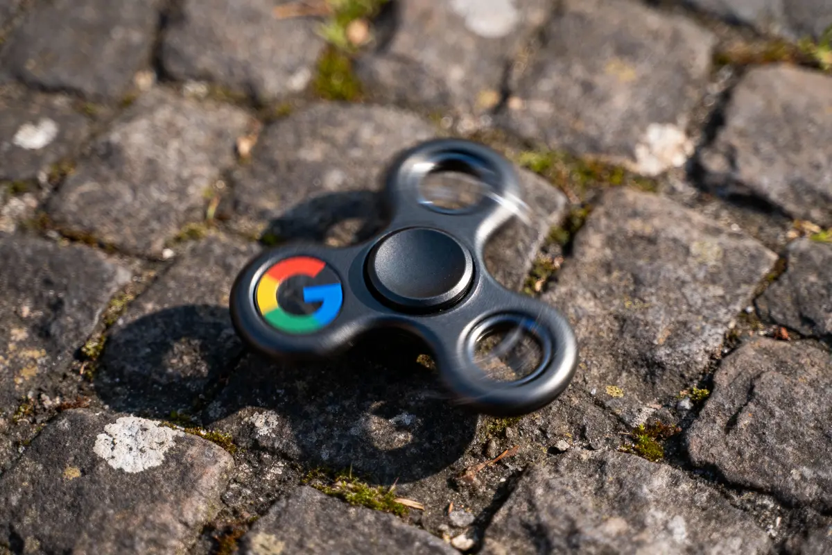Spinner noir avec logo coloré, posé sur pavés gris avec mousse verte.