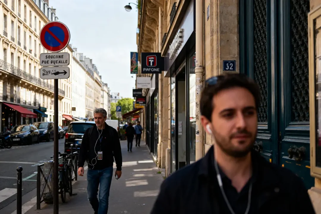 Homme marchant sur un trottoir parisien, écouteurs aux oreilles, devant boutiques et panneau de signalisation.