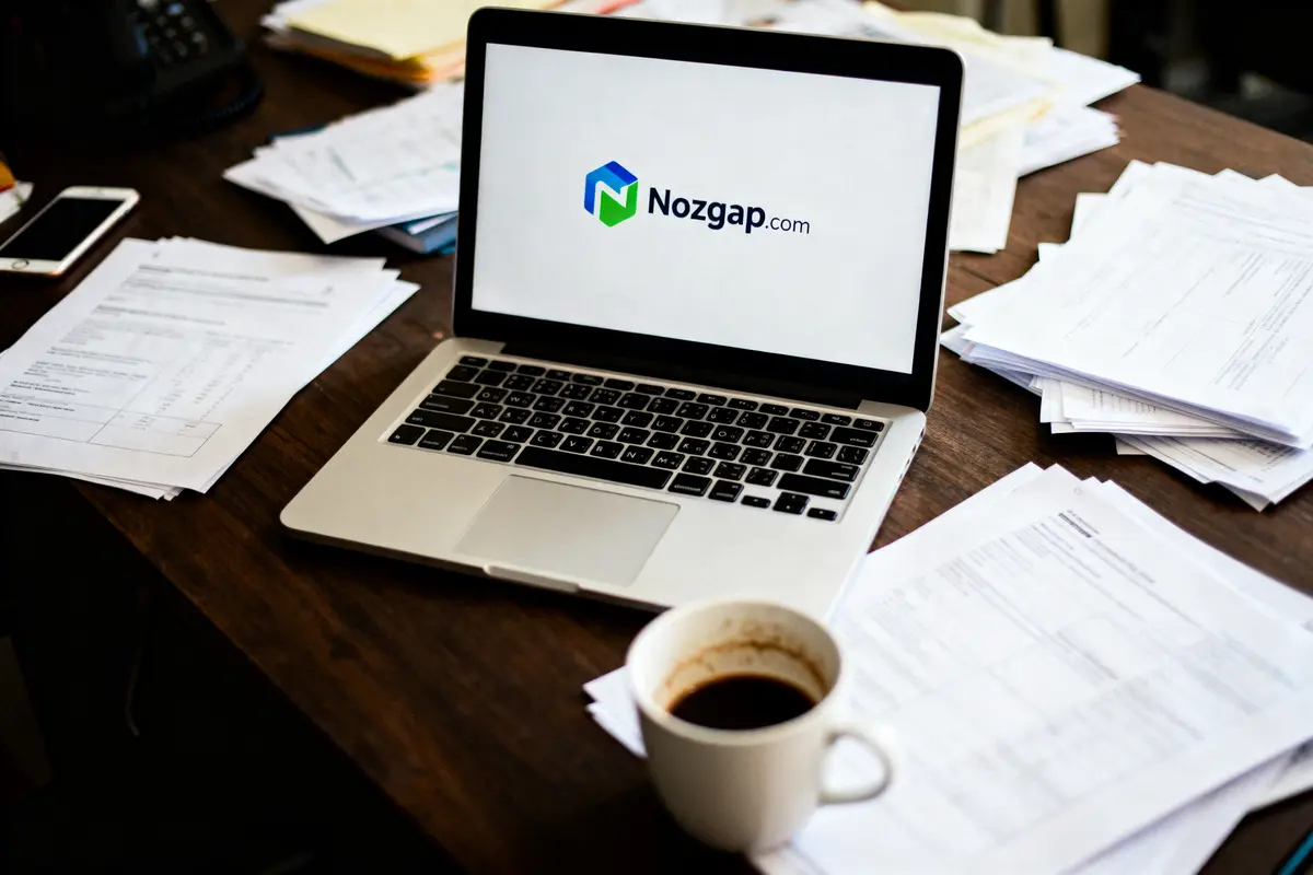Ordinateur portable affichant "Nozgap.com" sur bureau encombré de papiers, avec tasse de café.