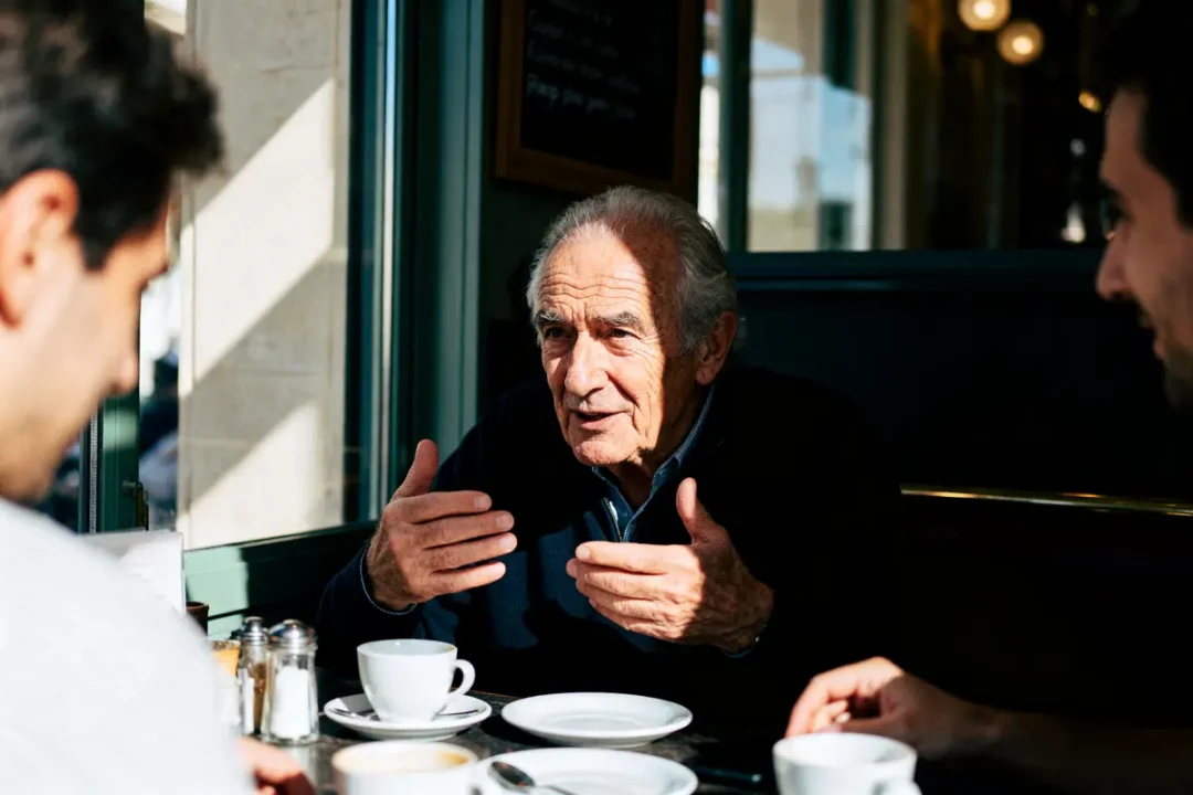 Un homme âgé discute avec deux personnes dans un café, gesticulant, autour de tasses de café.
