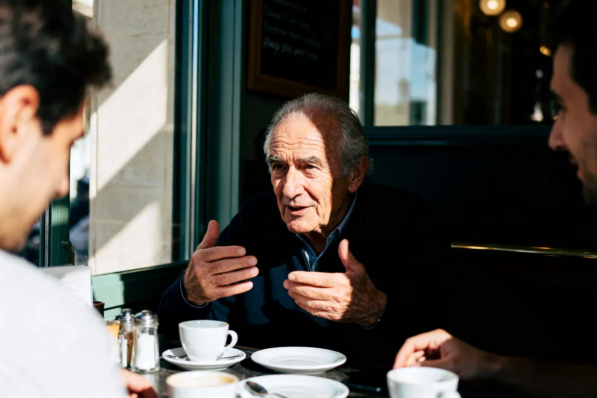 Un homme âgé discute avec deux personnes dans un café, gesticulant, autour de tasses de café.